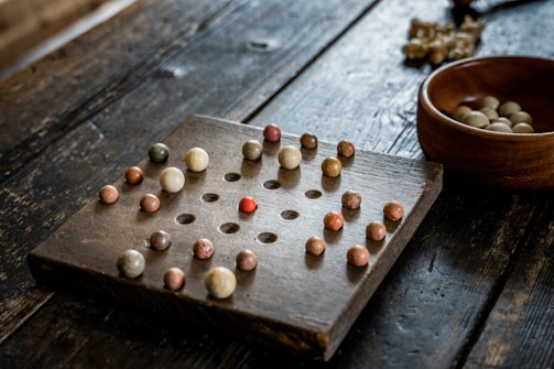 A wooden solitaire board with multiple small, colorful marbles positioned in holes. The board has a grid-like pattern, and there is an empty space in the center. A wooden bowl containing additional marbles is placed to the side on the rough-textured wooden surface.