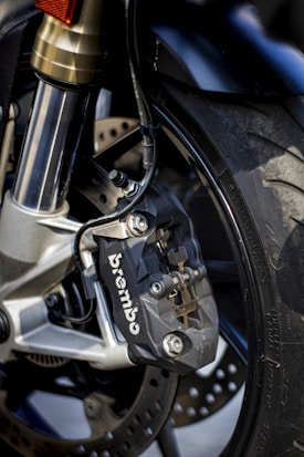 Close-up view of a motorcycle's front brake system, featuring a Brembo brake caliper attached to the suspension fork. The disc brake is visible behind the caliper with a pattern of holes for ventilation. The tire is partially visible, showing the textured tread pattern.