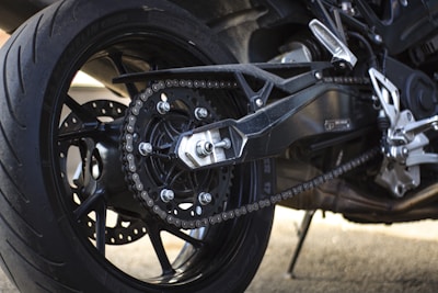 Close-up view of a motorcycle's rear wheel and drive chain, showcasing the intricate details of the sprocket and tire tread. The metal components are highlighted, displaying the engineering and design elements of the motorcycle.