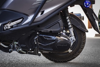 Close-up view of the rear wheel and suspension system of a scooter, highlighting the tire treads, shock absorber, and parts of the engine cover.
