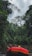 A red inflatable raft with paddles rests on the edge of a calm river surrounded by dense, lush green jungle foliage. Tall trees with a thick canopy rise into an overcast sky, creating a tranquil yet adventurous atmosphere.
