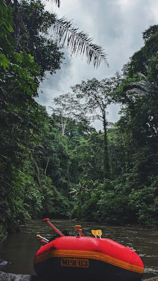 A red inflatable raft with paddles rests on the edge of a calm river surrounded by dense, lush green jungle foliage. Tall trees with a thick canopy rise into an overcast sky, creating a tranquil yet adventurous atmosphere.