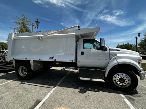 A large, white dump truck is parked in a lot. The truck is positioned to show its side profile with large wheels and a shiny exterior. There are markings and labels on the truck's body, and a lamppost and trees are visible in the background under a blue sky with some clouds.