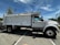 A large, white dump truck is parked in a lot. The truck is positioned to show its side profile with large wheels and a shiny exterior. There are markings and labels on the truck's body, and a lamppost and trees are visible in the background under a blue sky with some clouds.
