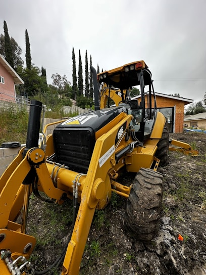 A large yellow backhoe is positioned on a muddy, grassy terrain with its front loader and large tires visible. In the background, there are tall evergreen trees and a small house with a peach-colored facade. The sky is overcast, adding a gray hue to the surroundings. A small building with an orange-brown facade is also visible nearby.