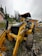 A large yellow backhoe is positioned on a muddy, grassy terrain with its front loader and large tires visible. In the background, there are tall evergreen trees and a small house with a peach-colored facade. The sky is overcast, adding a gray hue to the surroundings. A small building with an orange-brown facade is also visible nearby.