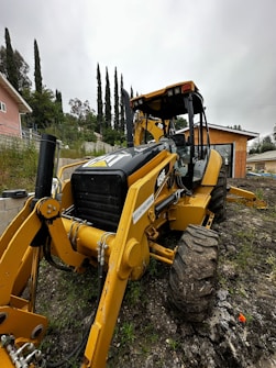 A large yellow backhoe is positioned on a muddy, grassy terrain with its front loader and large tires visible. In the background, there are tall evergreen trees and a small house with a peach-colored facade. The sky is overcast, adding a gray hue to the surroundings. A small building with an orange-brown facade is also visible nearby.