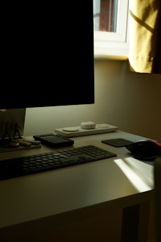 A tidy workspace with a black monitor, keyboard, and a calculator on a white desk. There are small decorative figurines beside the monitor and a wireless charging pad with two small devices on it. Sunlight is streaming through a window, casting a shadow across the desk.