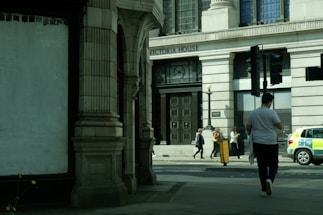 A street view featuring a large building labeled 'Victoria House' with ornate architectural features. Several people are walking on the sidewalk in front of the building, including one person in a white shirt walking away from the camera. A police car is parked on the side of the road.