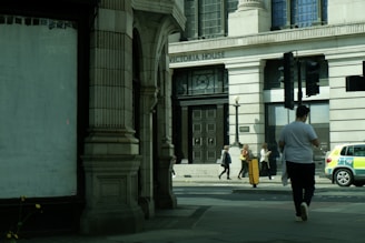 A street view featuring a large building labeled 'Victoria House' with ornate architectural features. Several people are walking on the sidewalk in front of the building, including one person in a white shirt walking away from the camera. A police car is parked on the side of the road.