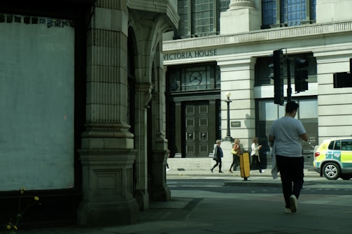 A street view featuring a large building labeled 'Victoria House' with ornate architectural features. Several people are walking on the sidewalk in front of the building, including one person in a white shirt walking away from the camera. A police car is parked on the side of the road.