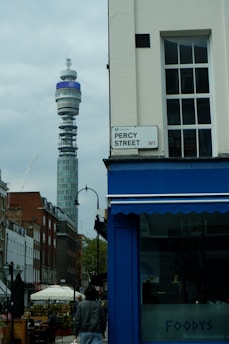 A bustling street scene in an urban area features a prominent telecommunications tower in the background. The foreground includes a street sign for Percy Street and a blue storefront labeled 'FOODYS'. Several buildings line the street, and a person is walking along the sidewalk. There are outdoor seating areas with umbrellas and visible greenery.
