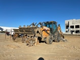 A construction site featuring a heavy-duty backhoe loader parked on sandy ground. The machine is predominantly yellow with some parts covered in dirt. In the background, there is an unfinished building structure and some vehicles, including a truck. The sky is clear and blue, suggesting a sunny day.