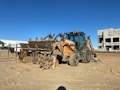A construction site featuring a heavy-duty backhoe loader parked on sandy ground. The machine is predominantly yellow with some parts covered in dirt. In the background, there is an unfinished building structure and some vehicles, including a truck. The sky is clear and blue, suggesting a sunny day.