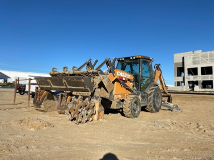 A construction site featuring a heavy-duty backhoe loader parked on sandy ground. The machine is predominantly yellow with some parts covered in dirt. In the background, there is an unfinished building structure and some vehicles, including a truck. The sky is clear and blue, suggesting a sunny day.