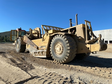 A large, heavy-duty construction vehicle is parked on a dirt construction site. The vehicle has enormous tires and is covered in dust and dirt. The background features a clear blue sky and a partially constructed building.