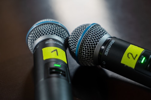 Two black and silver microphones with a mesh grille, each identified by a yellow label with the numbers 1 and 2. They are positioned on a dark surface, with blue bands around the grills and a small green light indicating power status.
