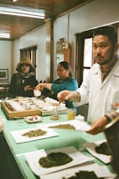 A group of people engaged in a tea tasting session in a cozy room with wooden interiors. Several small cups and loose tea leaves are arranged on a green table. One person is pouring liquid into a cup while another person is looking towards the camera. Another individual is in the foreground, also interacting with the tea setup.