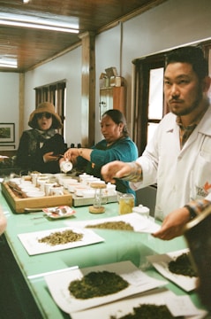 A group of people engaged in a tea tasting session in a cozy room with wooden interiors. Several small cups and loose tea leaves are arranged on a green table. One person is pouring liquid into a cup while another person is looking towards the camera. Another individual is in the foreground, also interacting with the tea setup.