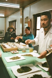 A group of people engaged in a tea tasting session in a cozy room with wooden interiors. Several small cups and loose tea leaves are arranged on a green table. One person is pouring liquid into a cup while another person is looking towards the camera. Another individual is in the foreground, also interacting with the tea setup.