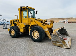A large yellow front loader with the number 950B displayed on the side is parked on a gravel lot. The machinery has big thick tires and a sizeable bucket, which shows signs of wear and some soil residue. In the background, there are distant construction materials and a hilly landscape under an overcast sky.