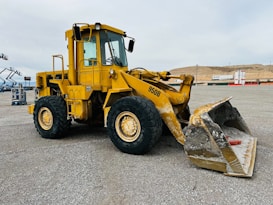 A large yellow front loader with the number 950B displayed on the side is parked on a gravel lot. The machinery has big thick tires and a sizeable bucket, which shows signs of wear and some soil residue. In the background, there are distant construction materials and a hilly landscape under an overcast sky.