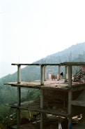 A partially constructed concrete building with exposed columns and floors is visible. Laundry is hanging on the upper floor, suggesting human presence or activity. The backdrop features a lush green forest and rolling hills, with a hazy sky creating a serene and somewhat isolated atmosphere.