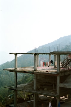 A partially constructed concrete building with exposed columns and floors is visible. Laundry is hanging on the upper floor, suggesting human presence or activity. The backdrop features a lush green forest and rolling hills, with a hazy sky creating a serene and somewhat isolated atmosphere.