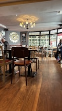 A cozy, warmly-lit cafe interior with wooden flooring and a variety of chairs and tables. A chandelier hangs from the ceiling, casting soft light, and ornate mirror decorations adorn the walls. Large windows allow natural light to stream in, creating a welcoming ambiance. A few patrons are seated, suggesting a relaxed, ambient atmosphere.