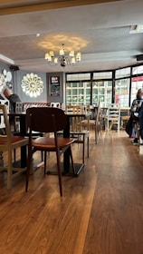 A cozy, warmly-lit cafe interior with wooden flooring and a variety of chairs and tables. A chandelier hangs from the ceiling, casting soft light, and ornate mirror decorations adorn the walls. Large windows allow natural light to stream in, creating a welcoming ambiance. A few patrons are seated, suggesting a relaxed, ambient atmosphere.