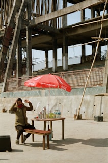 A person sits at a wooden table with a red Coca-Cola branded umbrella, set against the backdrop of an unfinished concrete structure. On the table are drinks, cups, and water bottles. The construction site appears to be empty, with wooden scaffolding and cement occupying the background.