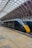 A sleek, modern train with a dark green and yellow exterior is stationed at a large indoor platform. The train's design is streamlined, and the platform is spacious with a tiled floor and a high arched ceiling made of metal and glass. Natural light filters through the ceiling, illuminating the scene.