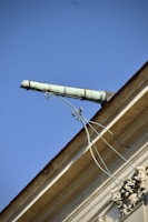 An ornate drainpipe extends from the corner of a building, supported by intricate wrought iron brackets. The architectural details include decorative elements at the base. The sky is bright and clear, providing a striking contrast to the building's structure.