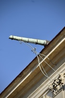 An ornate drainpipe extends from the corner of a building, supported by intricate wrought iron brackets. The architectural details include decorative elements at the base. The sky is bright and clear, providing a striking contrast to the building's structure.