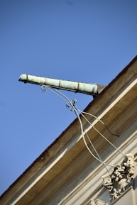 An ornate drainpipe extends from the corner of a building, supported by intricate wrought iron brackets. The architectural details include decorative elements at the base. The sky is bright and clear, providing a striking contrast to the building's structure.
