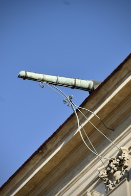 An ornate drainpipe extends from the corner of a building, supported by intricate wrought iron brackets. The architectural details include decorative elements at the base. The sky is bright and clear, providing a striking contrast to the building's structure.