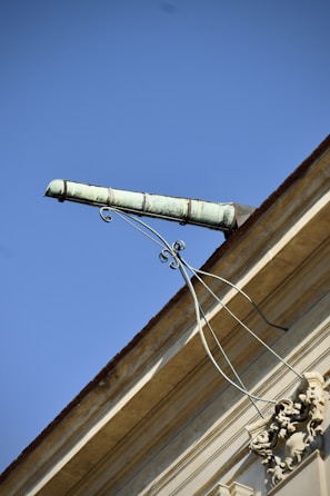 An ornate drainpipe extends from the corner of a building, supported by intricate wrought iron brackets. The architectural details include decorative elements at the base. The sky is bright and clear, providing a striking contrast to the building's structure.