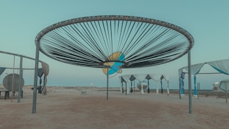 A large circular installation with a radial pattern of dark lines stretches across the sky. Below it, several structures with draped fabric canopies are arranged on a sandy surface, evoking a minimalist yet artistic environment. The background features a barren landscape under a clear sky, suggesting a tranquil, desolate setting.