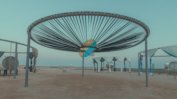 A large circular installation with a radial pattern of dark lines stretches across the sky. Below it, several structures with draped fabric canopies are arranged on a sandy surface, evoking a minimalist yet artistic environment. The background features a barren landscape under a clear sky, suggesting a tranquil, desolate setting.