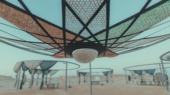 A series of large, circular canopy structures with intricate lattice patterns are seen in an arid desert landscape. Several suspended nets and coverings hang from metal frames, creating shaded areas underneath. The scene has a quiet and open atmosphere, with a clear pale blue sky above.