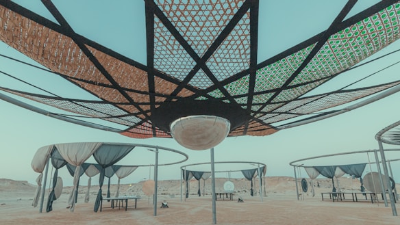 A series of large, circular canopy structures with intricate lattice patterns are seen in an arid desert landscape. Several suspended nets and coverings hang from metal frames, creating shaded areas underneath. The scene has a quiet and open atmosphere, with a clear pale blue sky above.