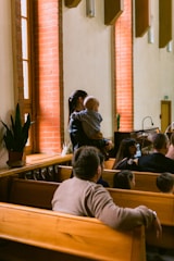 People are seated on wooden benches in what appears to be a church or chapel. A woman is standing while holding a baby near a brick wall with windows. Some plants are placed on a ledge near the window.