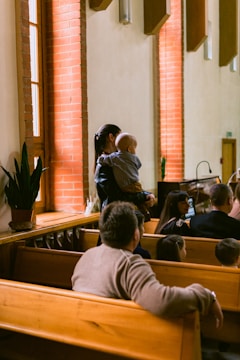 People are seated on wooden benches in what appears to be a church or chapel. A woman is standing while holding a baby near a brick wall with windows. Some plants are placed on a ledge near the window.