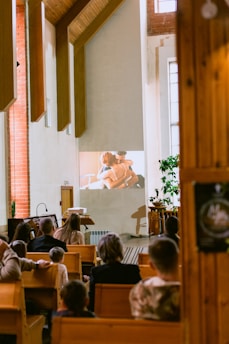 A group of people are seated in a room with wooden pews, watching a projected video on a wall. The setting appears to be a chapel or church with tall windows and wooden architectural features. The projection shows two people embracing, suggesting a scene from a film or presentation. The room is warmly lit, creating a serene atmosphere.