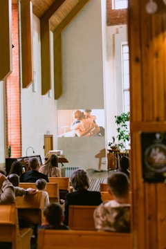 A group of people are seated in a room with wooden pews, watching a projected video on a wall. The setting appears to be a chapel or church with tall windows and wooden architectural features. The projection shows two people embracing, suggesting a scene from a film or presentation. The room is warmly lit, creating a serene atmosphere.