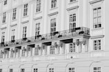 A detailed view of a classical building façade with large windows, ornate wrought-iron balconies, and decorative architectural elements such as corbels beneath the balcony. The building is rendered in shades of gray, indicating a black and white photograph.