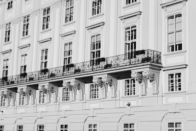 A detailed view of a classical building façade with large windows, ornate wrought-iron balconies, and decorative architectural elements such as corbels beneath the balcony. The building is rendered in shades of gray, indicating a black and white photograph.