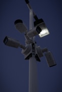 Several security cameras are mounted on a tall pole, with a bright light fixture attached nearby. The background is a dark evening sky.