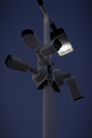 Several security cameras are mounted on a tall pole, with a bright light fixture attached nearby. The background is a dark evening sky.