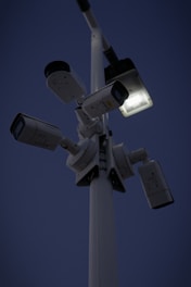 Several security cameras are mounted on a tall pole, with a bright light fixture attached nearby. The background is a dark evening sky.